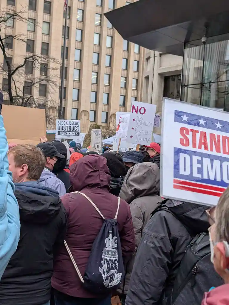 A crowd of protesters at an outdoor rally, many wearing jackets and hoodies, holding signs including one that reads 'NO…