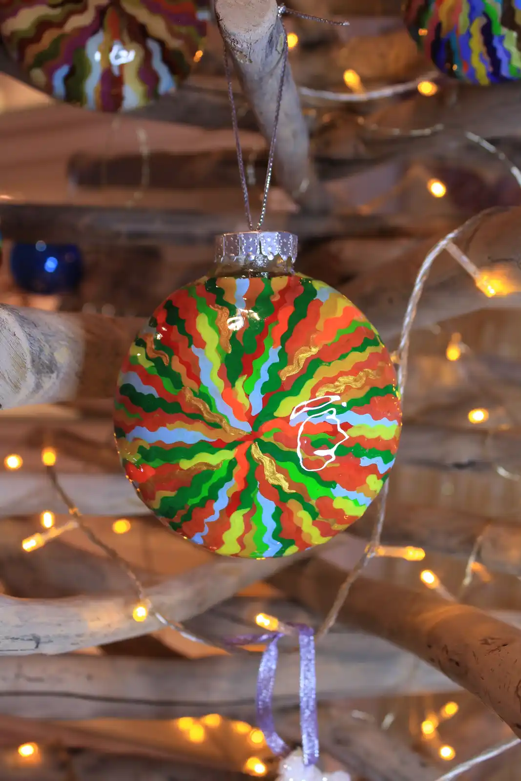 A close-up photo shows a colorful, hand-painted Christmas ornament with wavy stripes of red, green, yellow, blue, and gold…