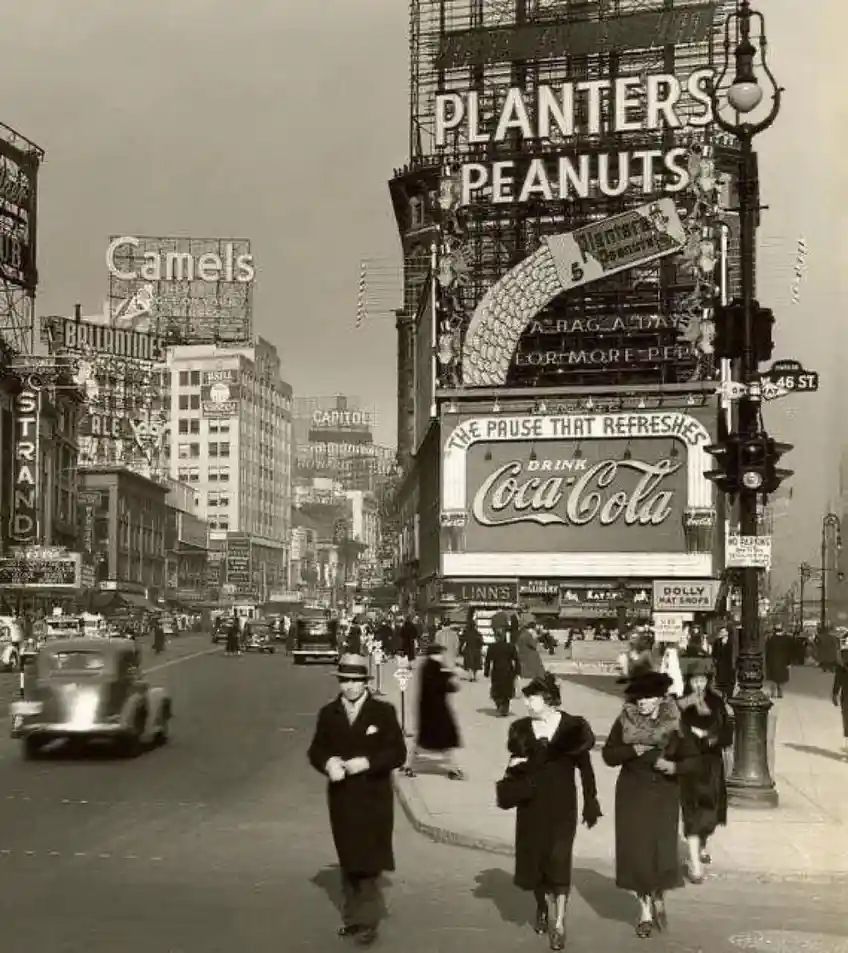 A vintage black-and-white photograph of a bustling 1930s New York City street, likely Times Square, filled with pedestrians…