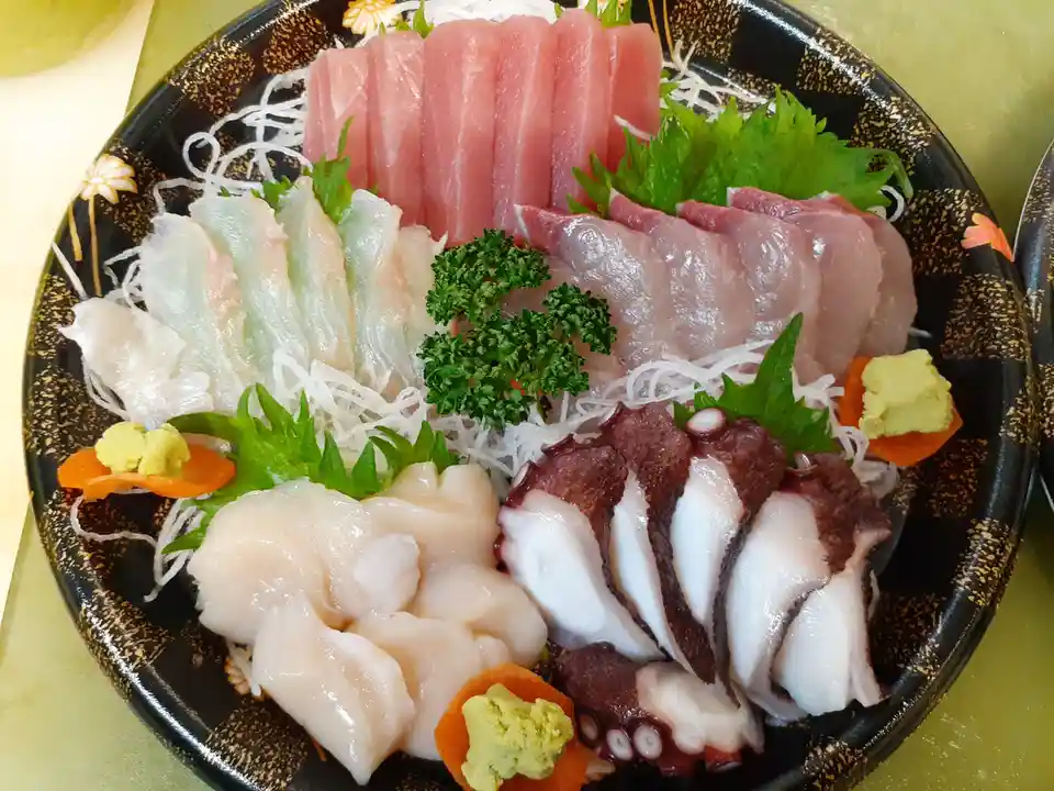 A close-up photo of a black ceramic plate filled with assorted raw seafood sashimi, including pink tuna, white fish…