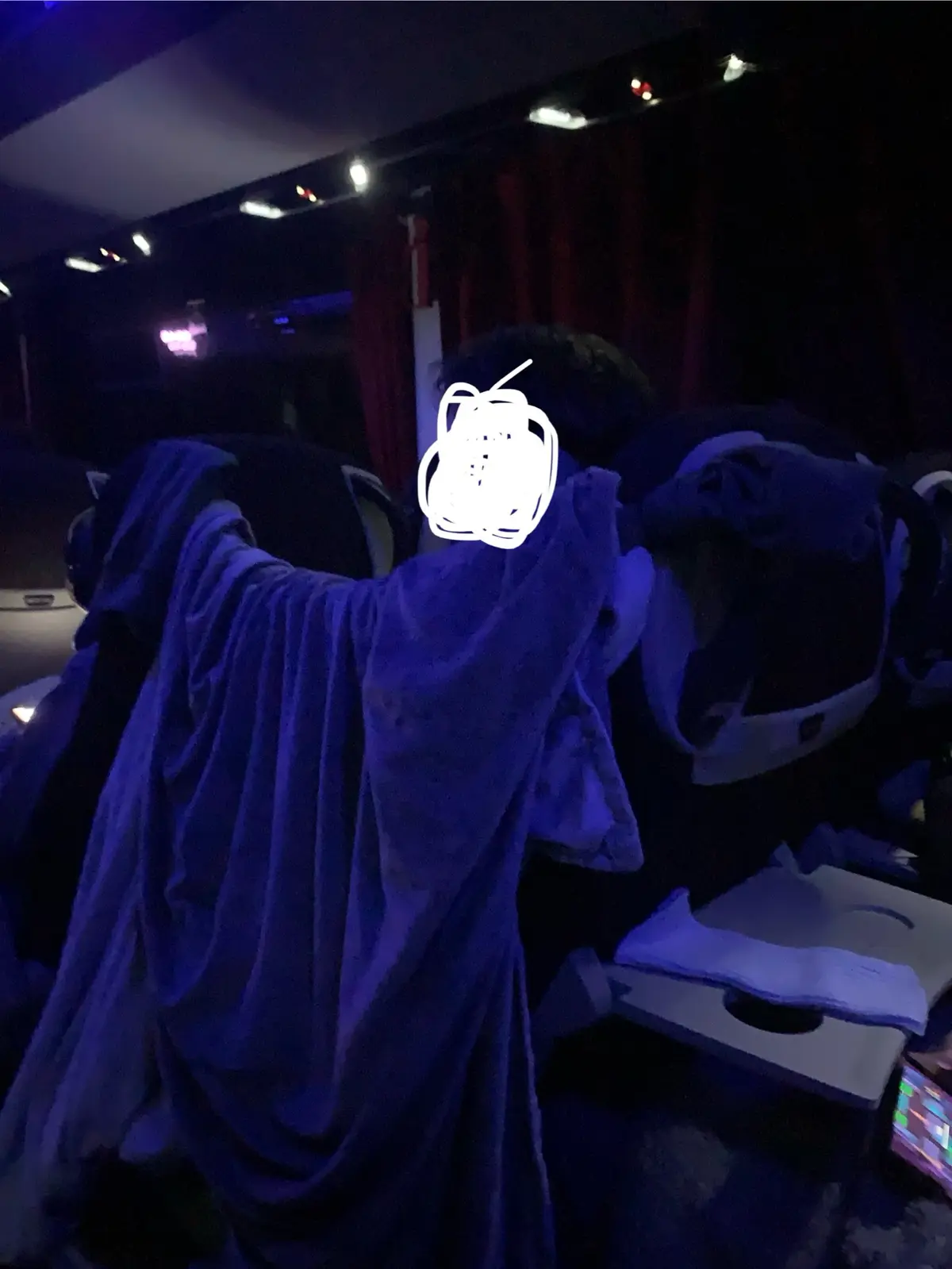 A person in a dimly lit room, possibly a theater or concert venue, is draped in a purple blanket while sitting in a chair…