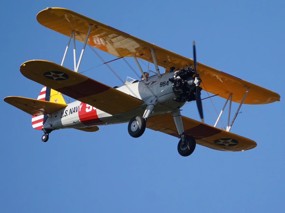 A vintage yellow and white U.S. Navy biplane with red accents and star insignia is flying against a clear blue sky. The…