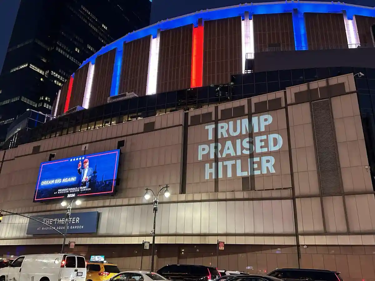 A nighttime photo of Madison Square Garden's exterior, featuring a large projection that reads 'TRUMP PRAISED HITLER' and a…