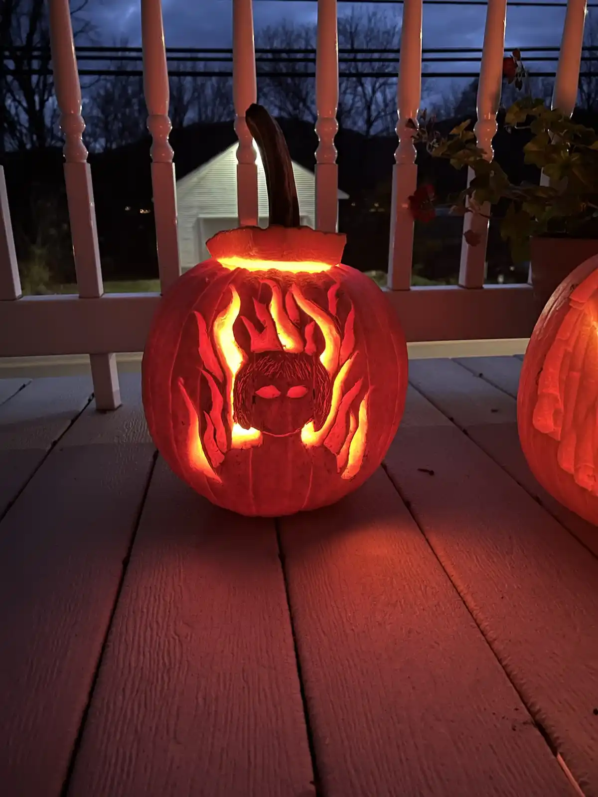 A carved Halloween pumpkin sits on a wooden deck at night, illuminated from within by a warm orange light. The pumpkin…