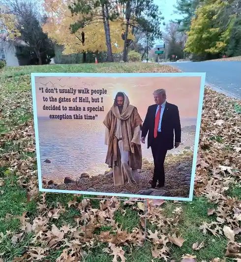 A yard sign placed in grass with fallen leaves shows a composite image of Jesus Christ and Donald Trump walking side by side…