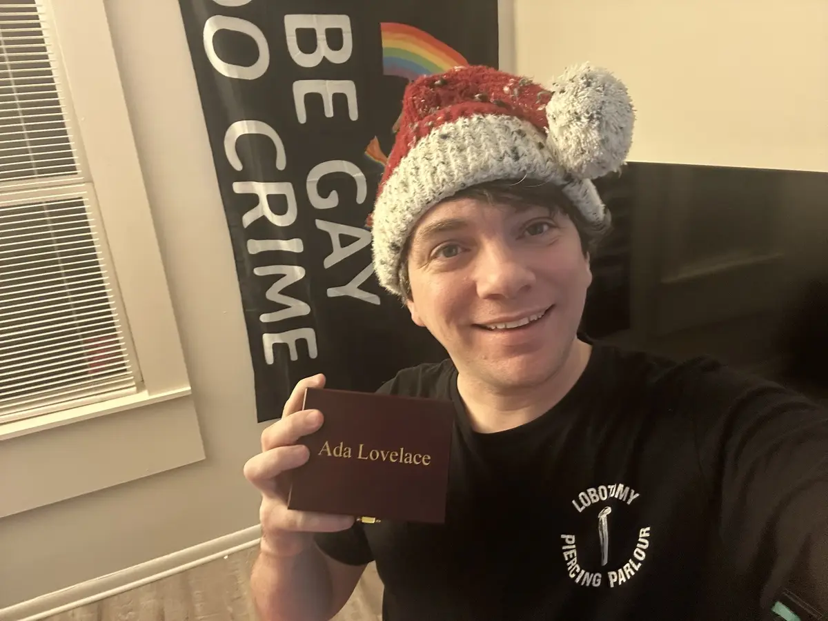 A person wearing a red and white knitted hat with fluffy ears smiles while holding up a small brown box labeled 'Ada…