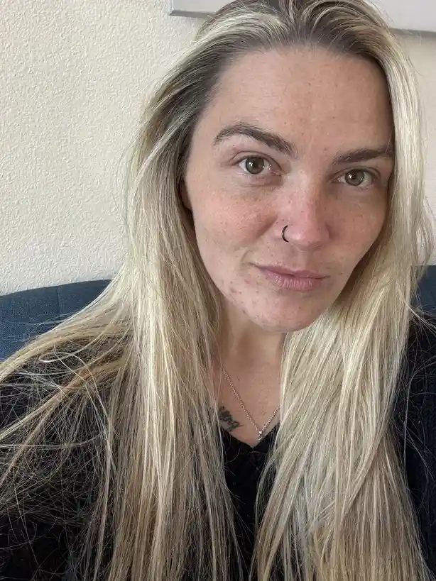 A close-up selfie of a woman with long blonde hair, freckles, and a nose ring, looking directly at the camera with a…