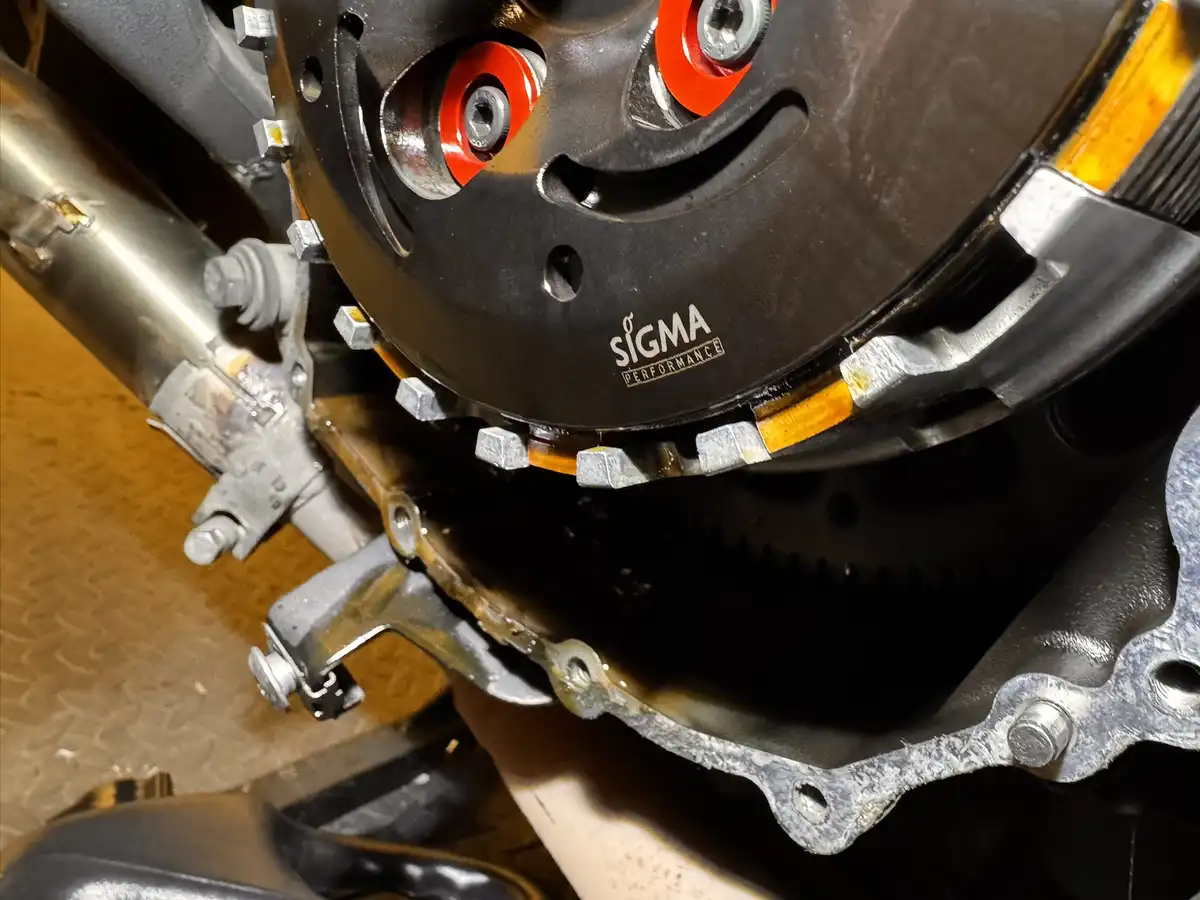 A close-up photo of a motorcycle's engine components, showing a black clutch cover with 'SIGMA PERFORMANCE' branding, red…