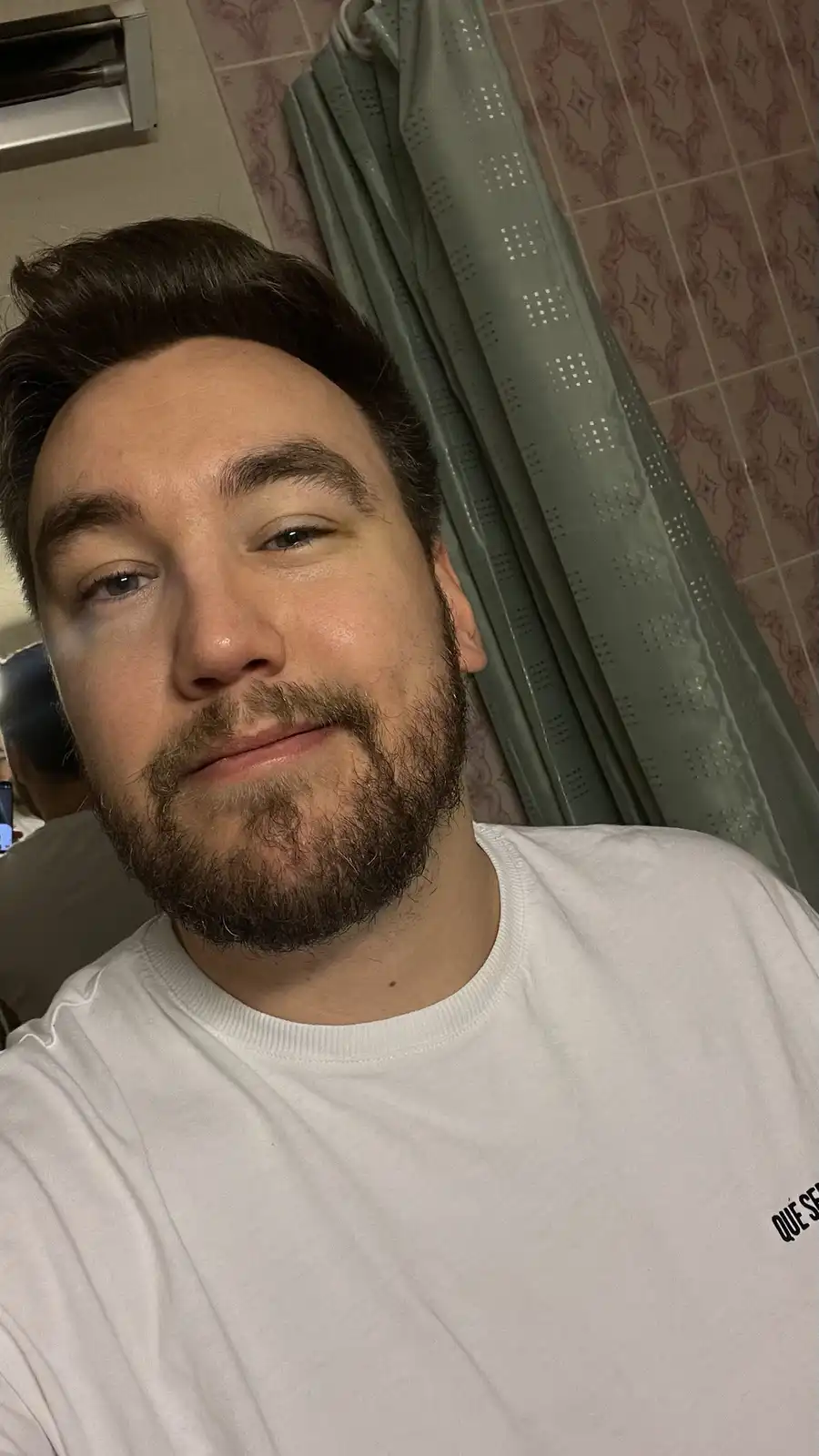 A selfie of a man with a beard and styled hair, wearing a white t-shirt, looking at the camera with a slight smirk. Behind…