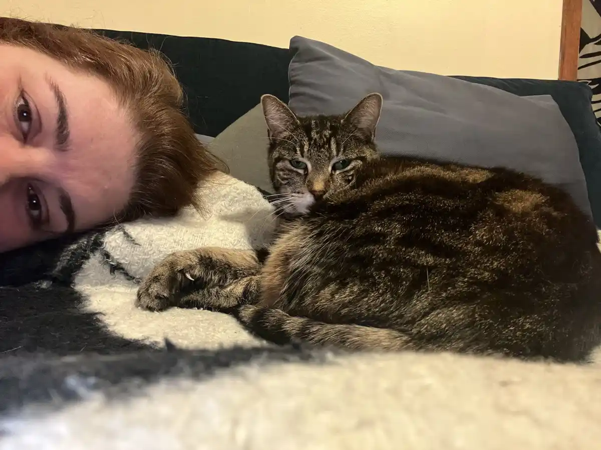A person with reddish-brown hair lies on a couch next to a tabby cat, both looking at the camera. The cat has a visibly…