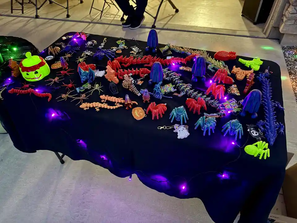 A table covered in a black cloth displays a wide variety of colorful, glowing Halloween-themed crafts including spiders…