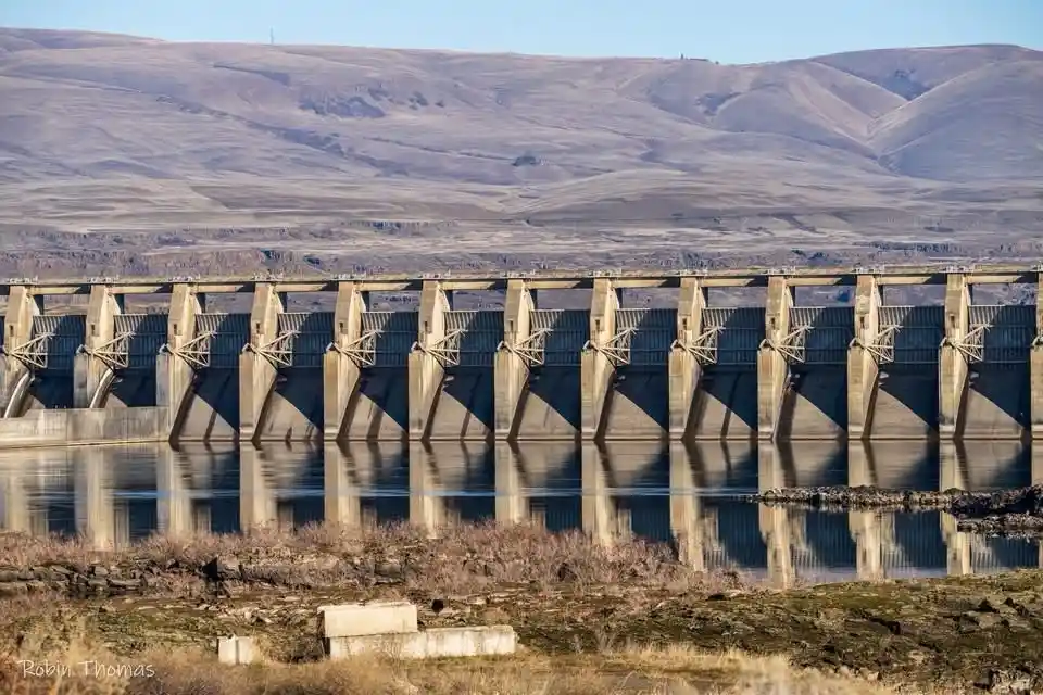 A wide shot of a large concrete dam with multiple vertical supports, spanning across a calm body of water that reflects its…