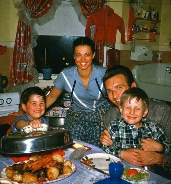 A vintage family photo shows a smiling woman, man, and two young boys gathered around a dining table laden with food…