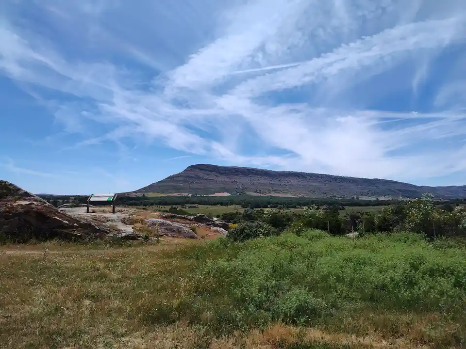 A scenic landscape photo shows a grassy field with large rocks in the foreground, a distant mountain range under a blue sky…