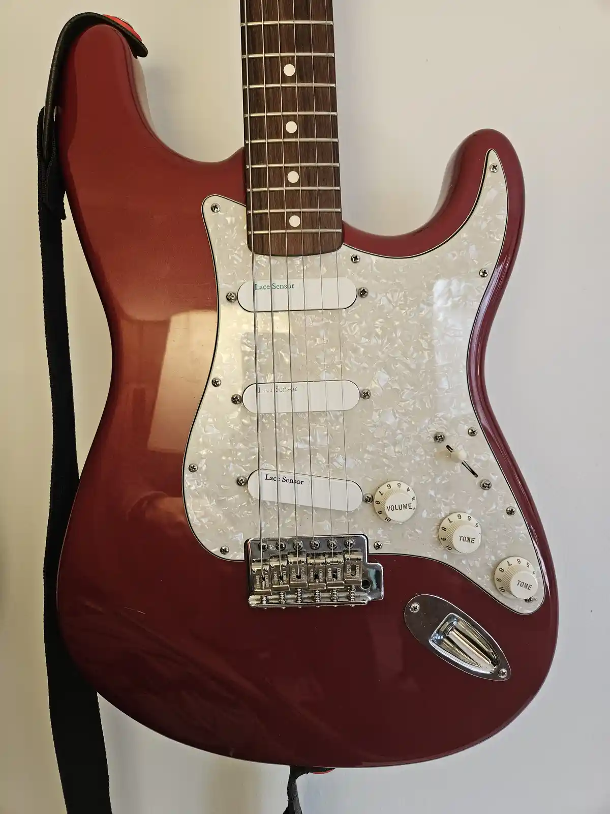 A red electric guitar with a white pickguard and three pickups, each labeled 'Lace Sensor', is shown against a plain wall…