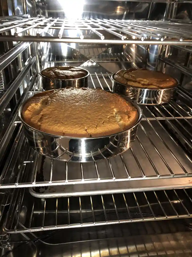 Three golden-brown baked cakes in metal pans sit on oven racks, with the front cake visibly cracked and risen unevenly…