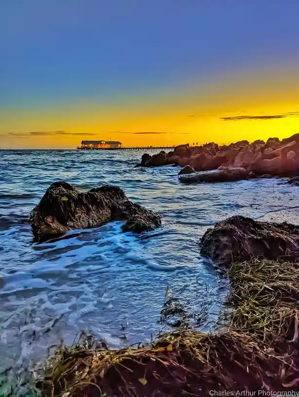 A vibrant sunset photo shows a pier with a lit building extending over the ocean, with waves crashing against rocks in the…