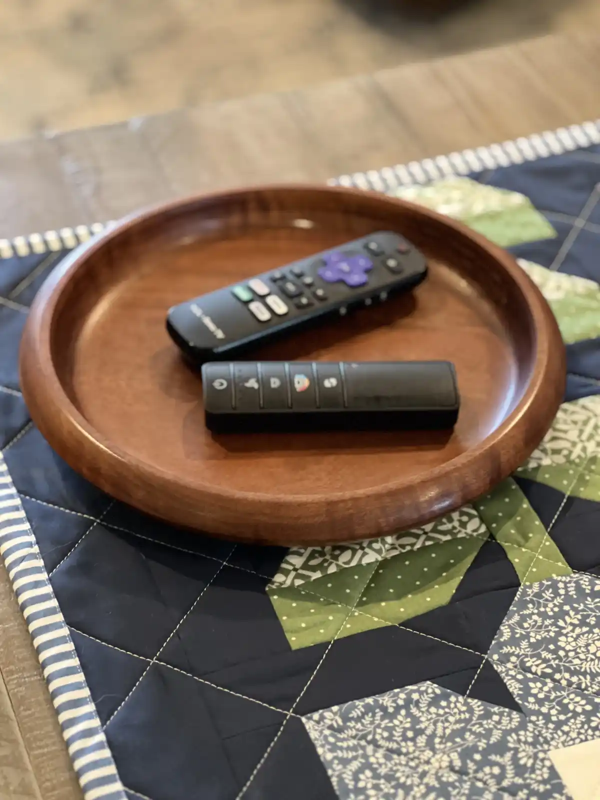 A wooden tray holds two remote controls on a patchwork quilt. One is a standard TV remote with a purple directional pad, the…