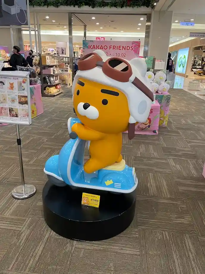 A large, yellow cartoon bear statue wearing a white pilot helmet with brown goggles is seated on a blue scooter display in a…