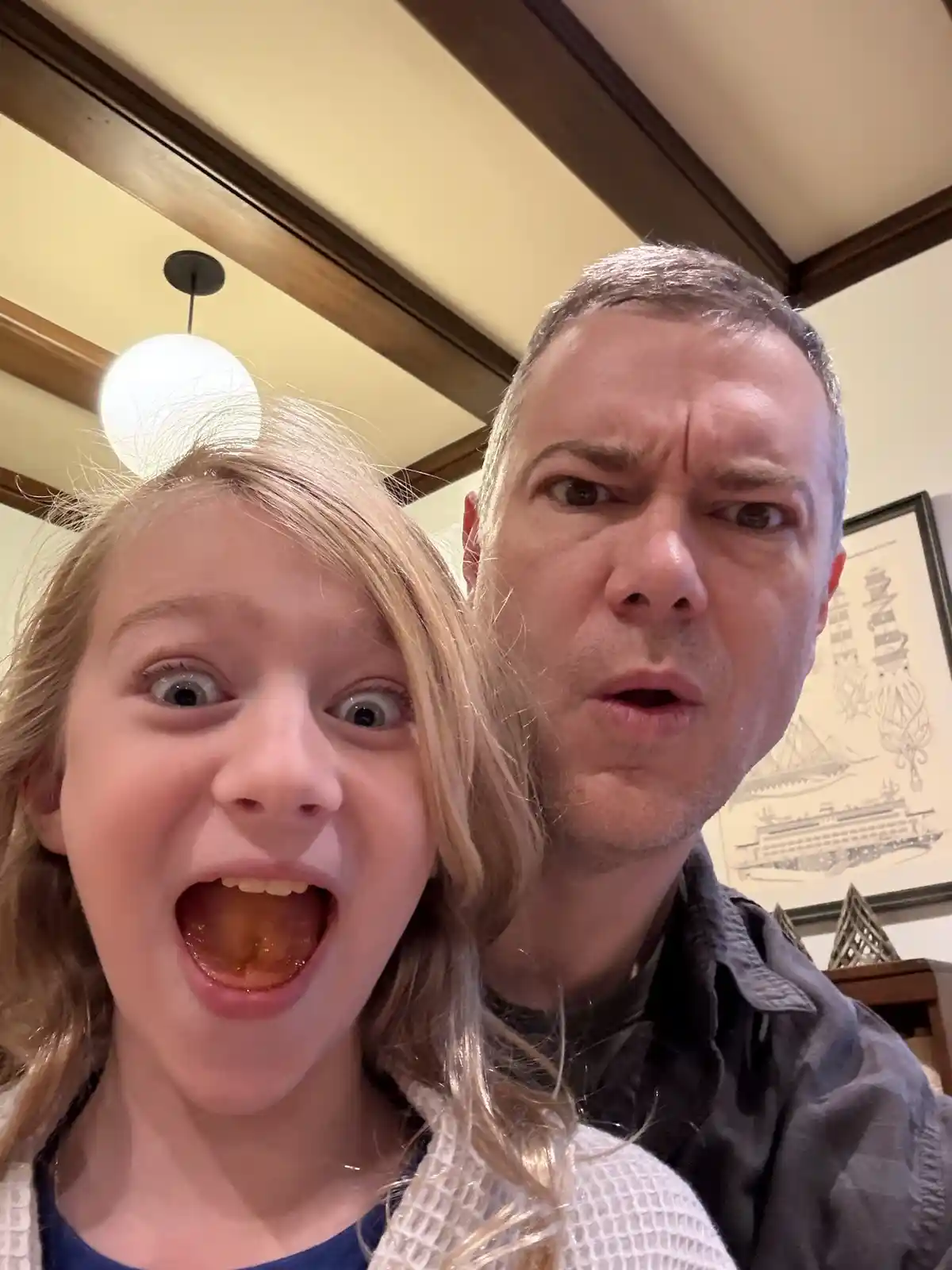 A selfie shows a young girl with wide eyes and an open mouth, revealing food residue, while a man beside her makes a…