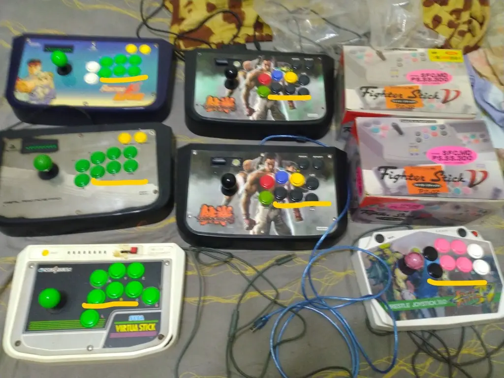 A collection of six different fighting game arcade sticks laid out on a bedspread, each with a yellow line drawn…