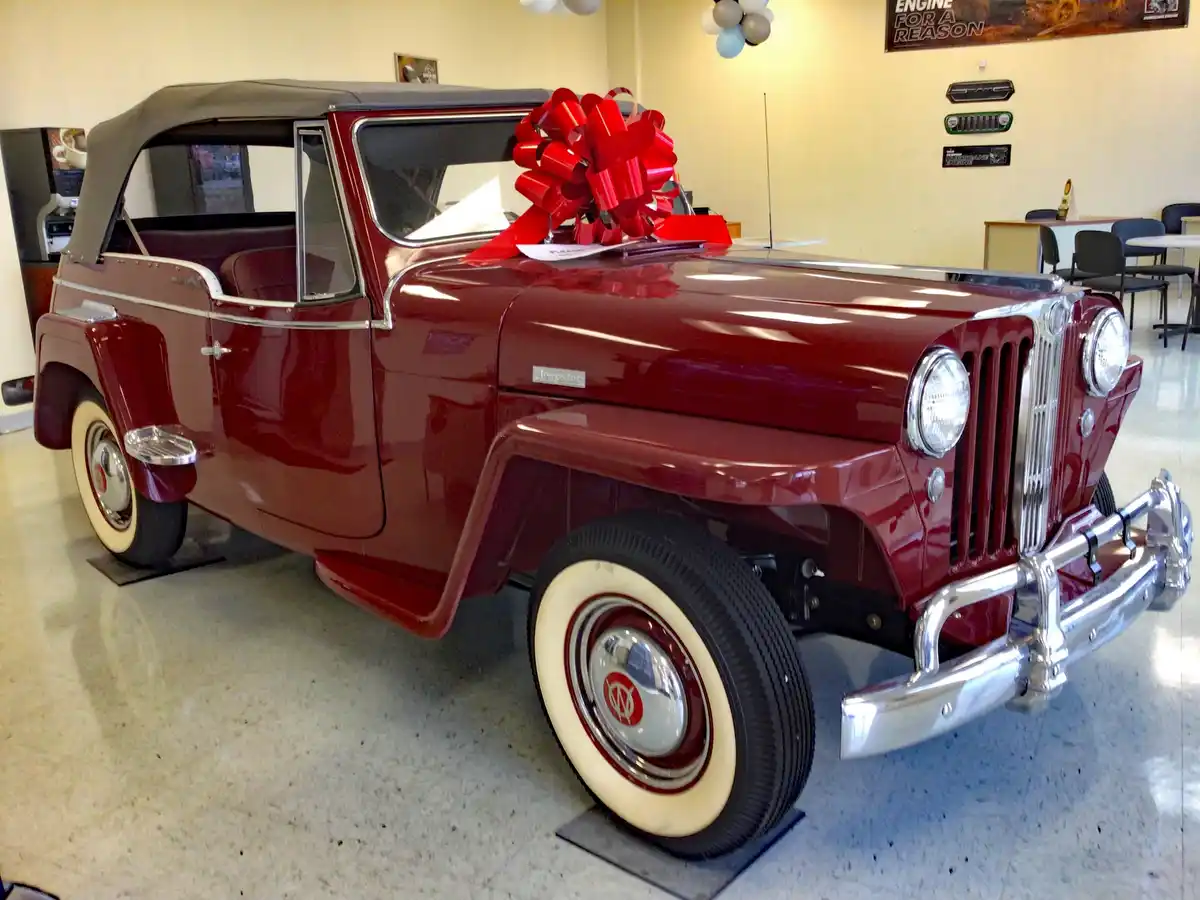 A shiny maroon vintage Jeepster convertible is displayed indoors on black platforms, adorned with a large red bow on its…