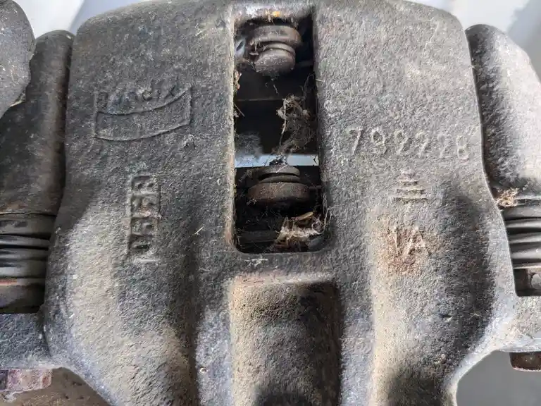 A close-up of a rusty, dirty automotive brake caliper with the Amazon logo and '792228' stamped on it, filled with cobwebs…
