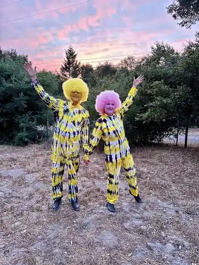 Two people stand outdoors at sunset wearing matching yellow, black, and white tie-dye jumpsuits and oversized afro wigs—one…