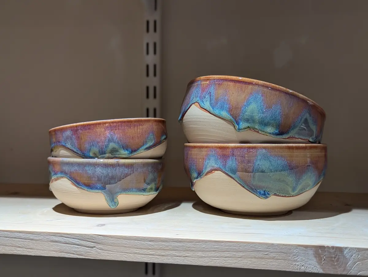 Two stacks of handmade ceramic bowls with glossy, colorful glaze that drips down their sides, sitting on a wooden shelf. The…