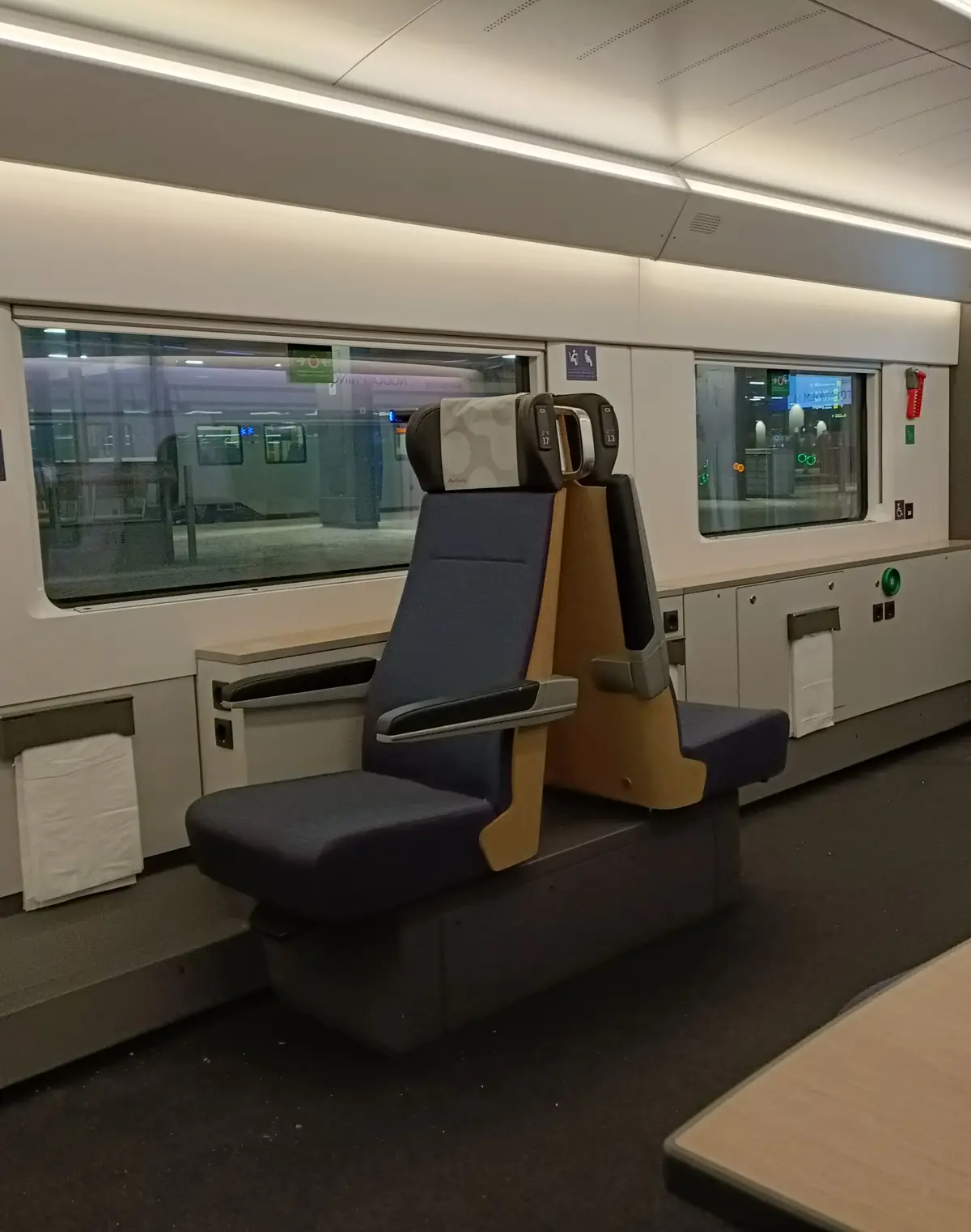 An empty, modern train seat with blue upholstery and wooden accents is shown inside a train car, facing a window with a…