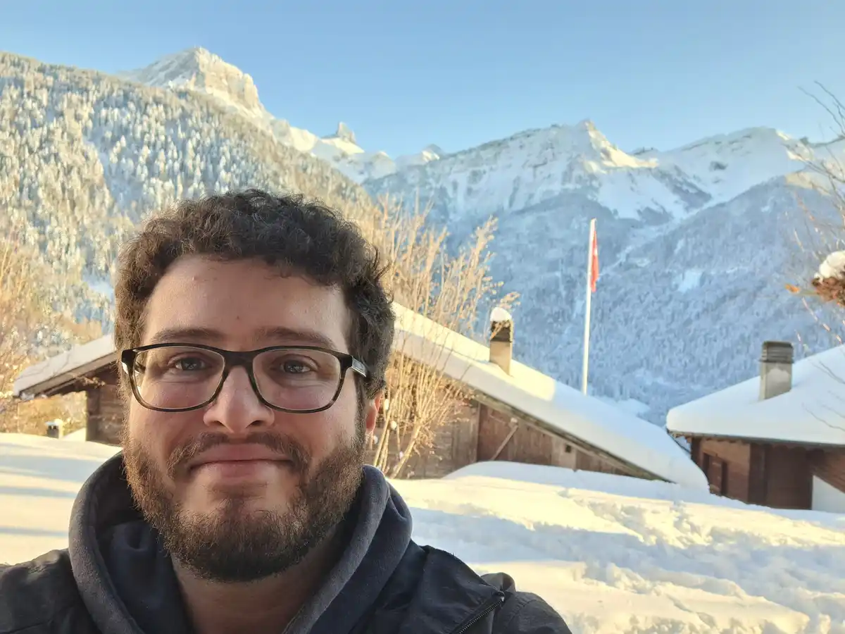 A man with glasses and a beard takes a selfie in front of snow-covered mountains and wooden chalets, smiling slightly. A…
