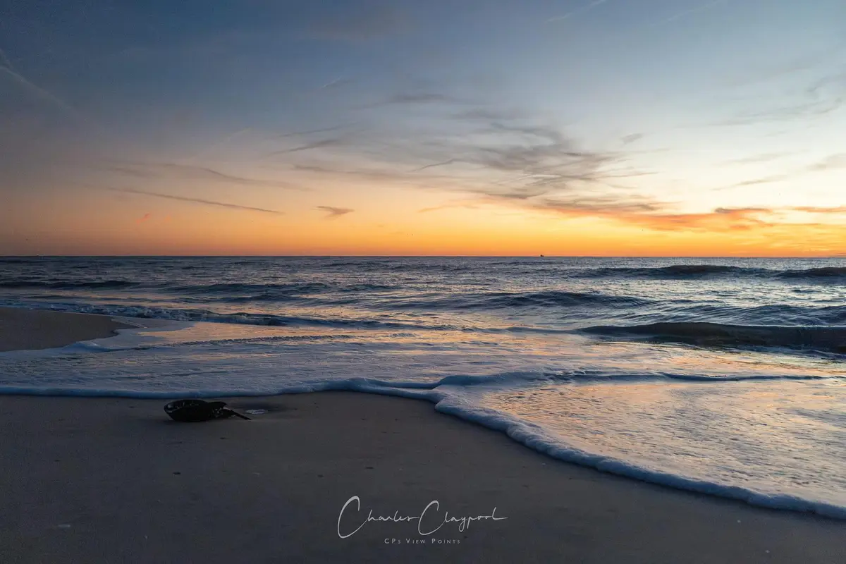 A serene beach sunset photo with gentle waves rolling onto the sand, a small dark object lies near the water's edge, and the…