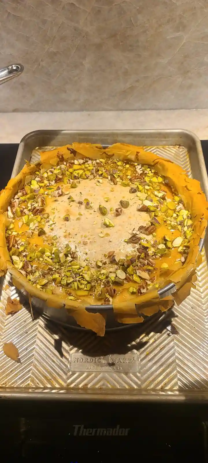 A close-up photo shows a round baklava dessert in a metal springform pan, sitting on a larger metal baking sheet. The…
