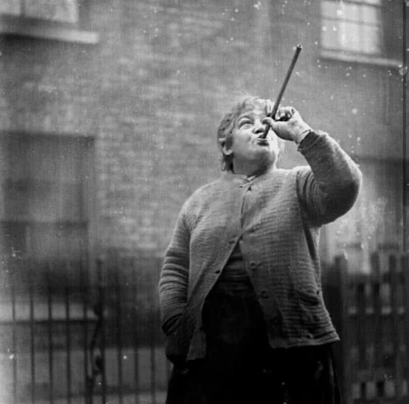 A black-and-white photo shows an older woman outdoors, holding a long stick or cane up to her mouth as if blowing into it…