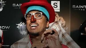 A man wearing a red hat, blue headband, and a red clown nose grimaces while holding a small object. He wears thick-rimmed…
