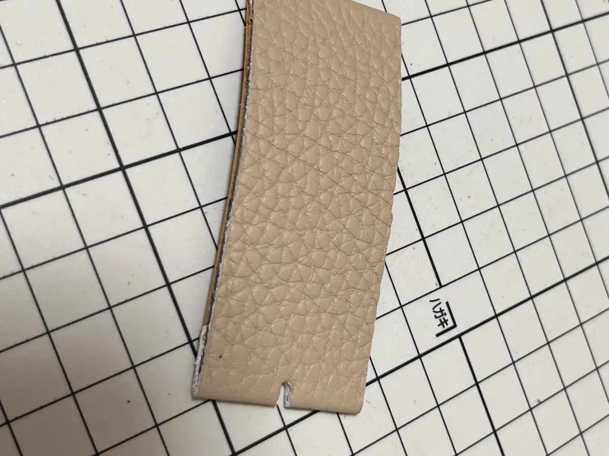 A beige textured leather strip with a torn edge lies diagonally on a white grid-patterned surface, with the Japanese…