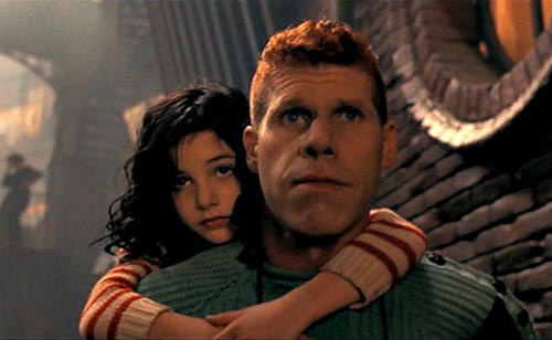 A man with red hair and a green sweater is being hugged from behind by a young girl with dark curly hair. He looks upward…
