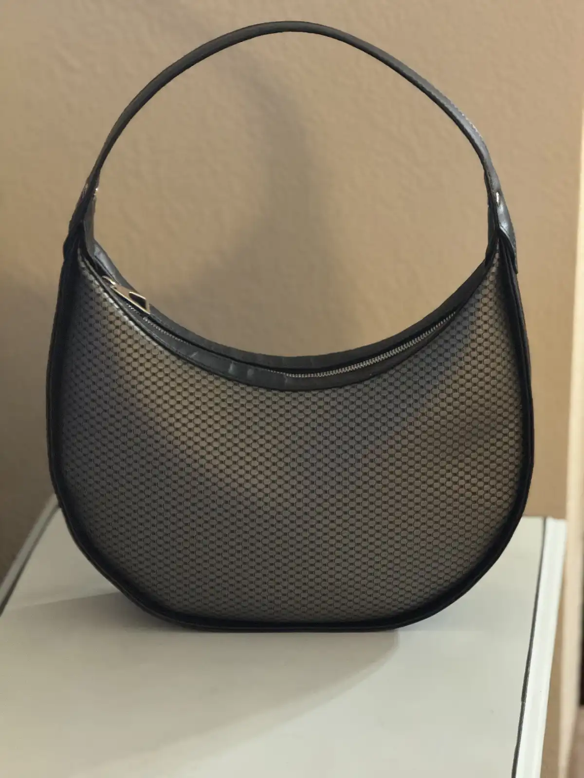 A black, semi-transparent mesh hobo-style handbag with a curved top handle sits on a white surface against a beige wall. The…