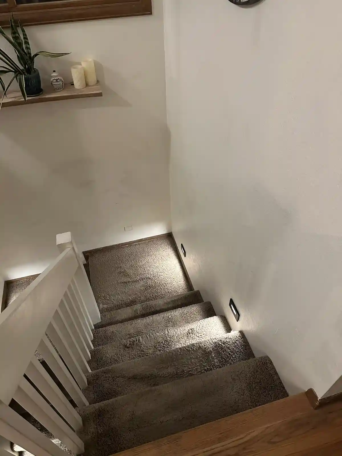 An overhead shot of a carpeted staircase with white railings, leading downward into a dimly lit hallway. On the wall above…