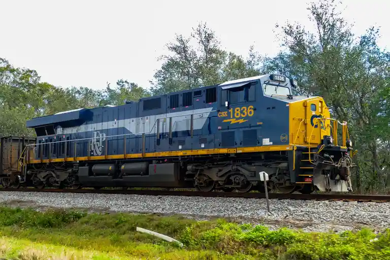 A blue and yellow CSX locomotive, number 1836, is shown on a railway track surrounded by trees and grass. The overlaid text…