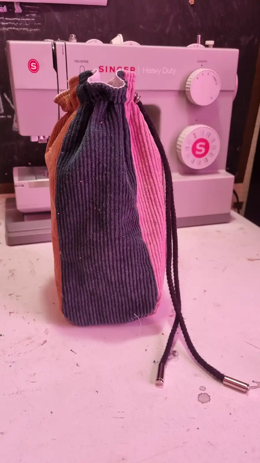A handmade drawstring bag, crafted from ribbed sweater fabric in orange, dark gray, and pink sections, sits on a work…
