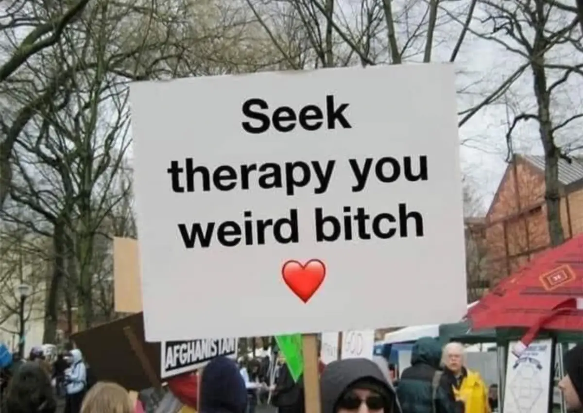 A white protest sign held outdoors among trees and other protesters displays the text 'Seek therapy you weird bitch' in bold…