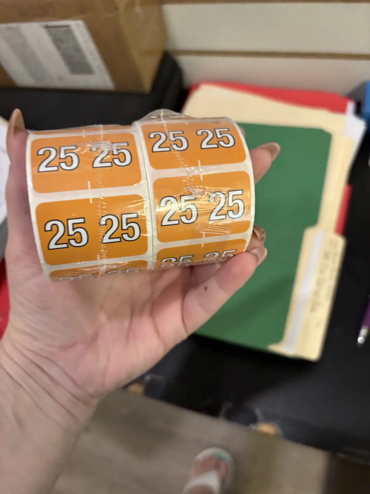 A hand holds a roll of orange adhesive labels, each printed with the number '25' twice in white. The background shows office…