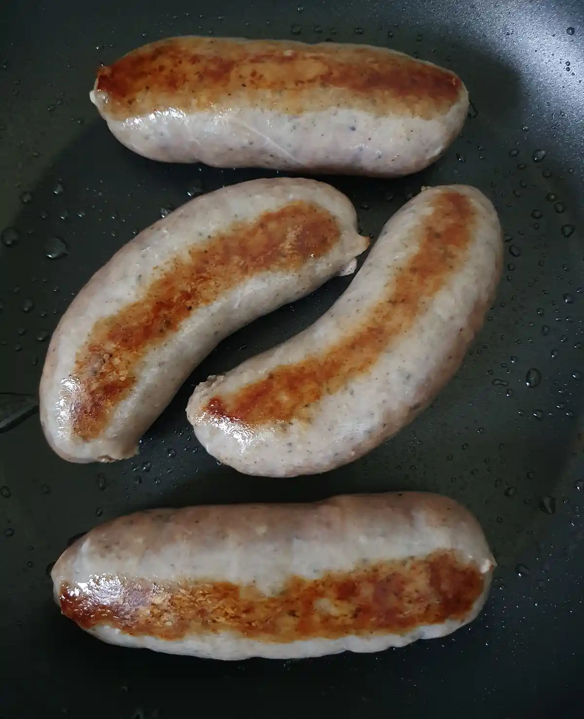 Four sausages are frying in a black pan, each showing distinct browned, seared stripes on their surfaces. The pan has…
