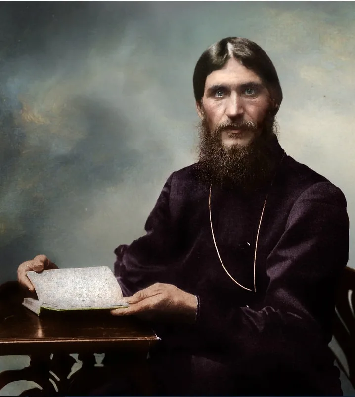A vintage portrait of a bearded man in dark religious robes, seated at a wooden table holding an open book. He stares…