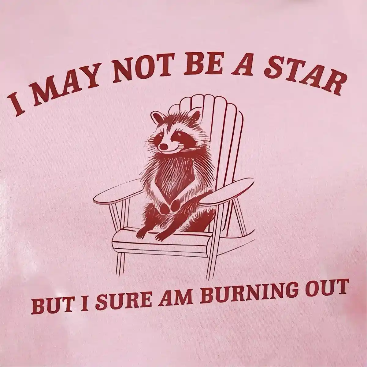 A pink t-shirt features a red illustration of a raccoon sitting in a wooden Adirondack chair. Above the raccoon, the text…