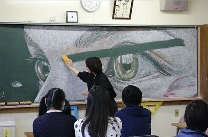 A teacher in a classroom is erasing a large, detailed chalk drawing of a human eye on a blackboard, while students sit…