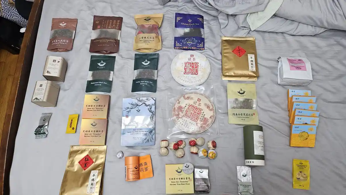 An overhead shot displays a large, diverse collection of tea products—bags, tins, cakes, and small packets—arranged neatly…