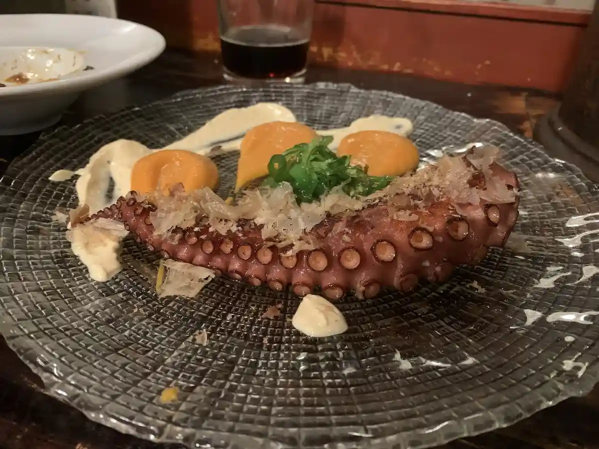 A close-up photo of a plated grilled octopus tentacle on a textured glass dish, topped with shredded bonito flakes, green…