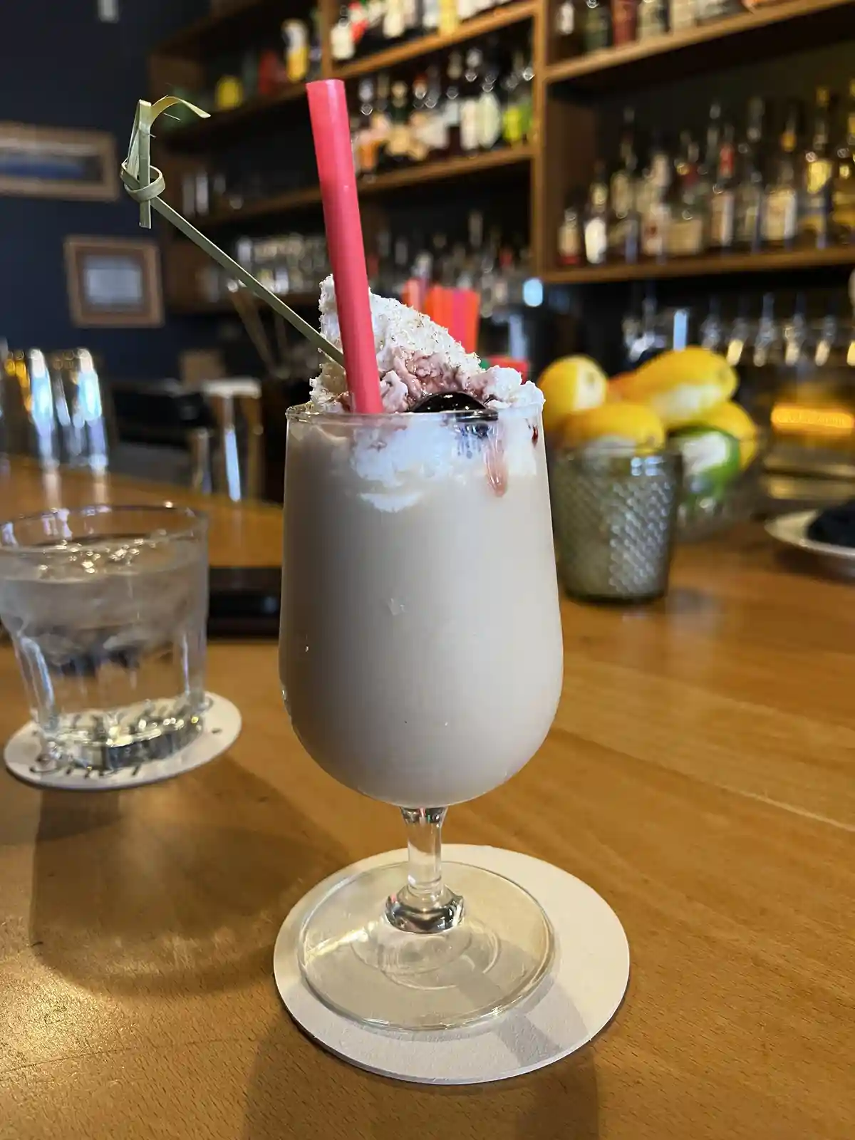A creamy milkshake in a stemmed glass sits on a coaster on a wooden bar, topped with whipped cream, a cherry, and a pink…